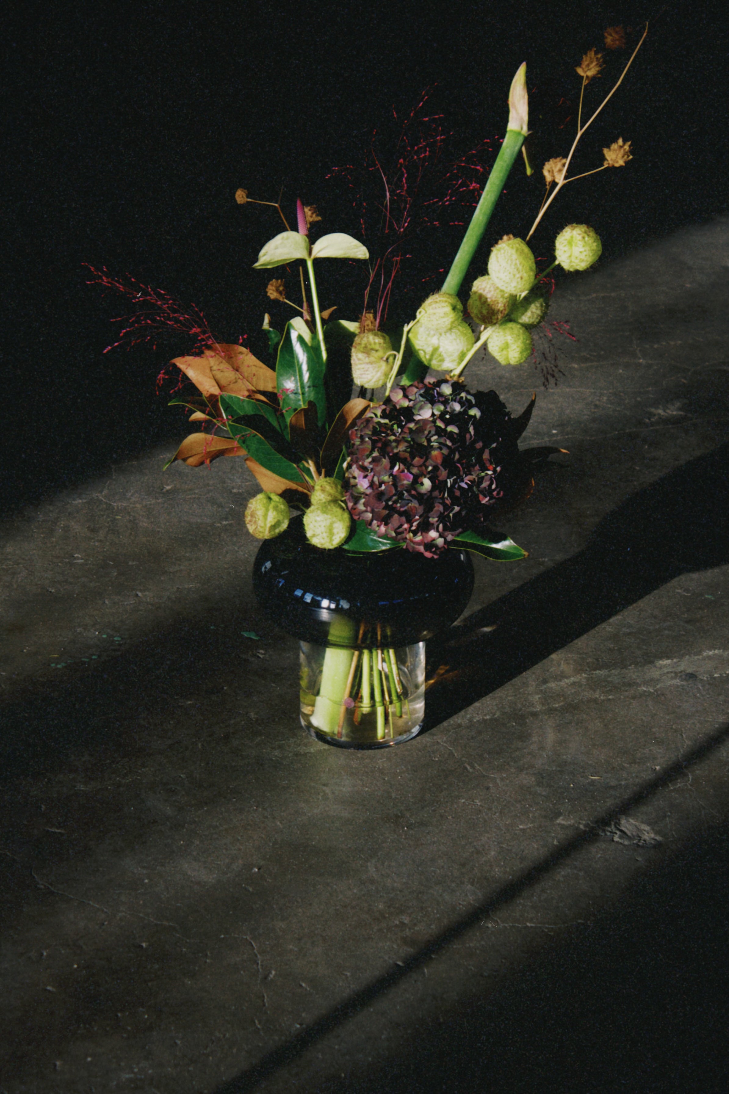 Smoked-glass vase with dried stems on a fabric-covered table, backlit by a large window.